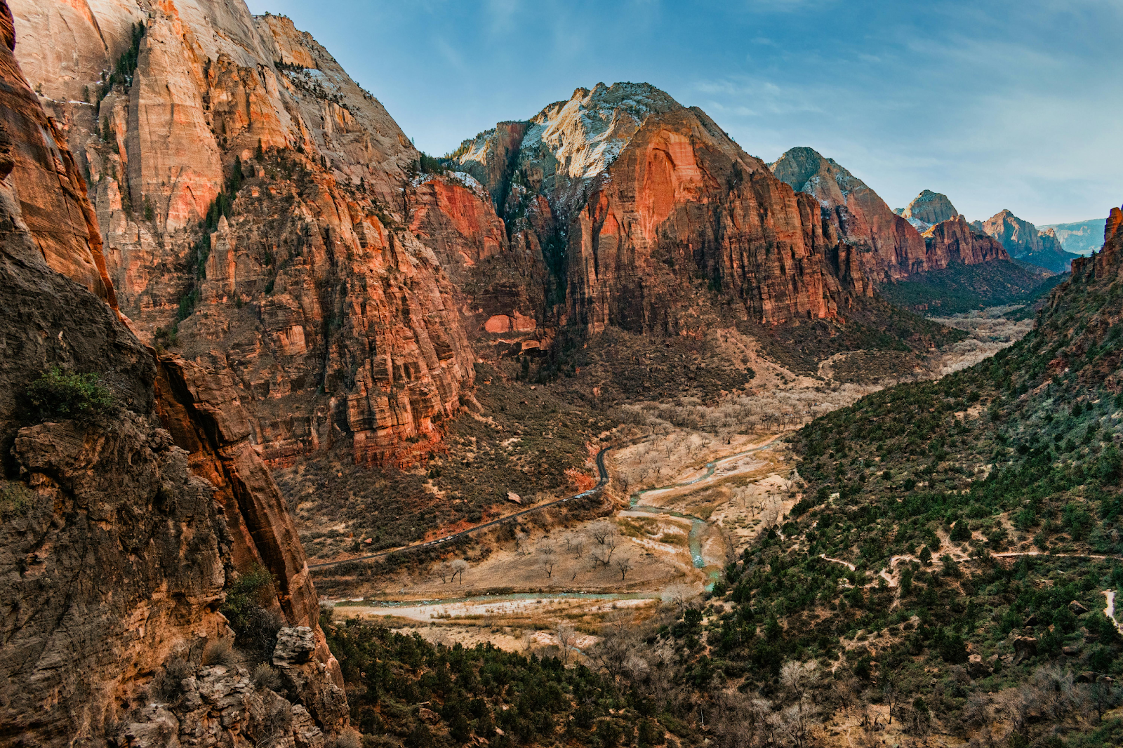 A wide-angle view of a deep canyon with towering, reddish-orange rock faces. A winding river and a small road snake through the valley floor, which is dotted with green trees and dry brush.