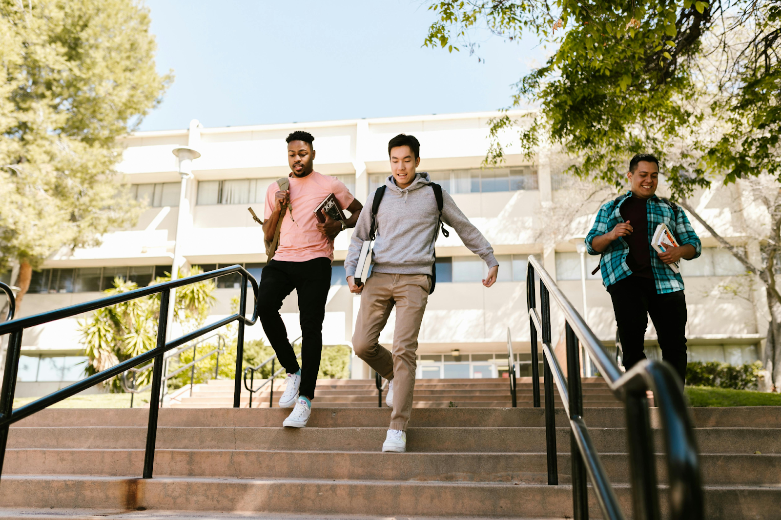 Three young men carrying books and backpacks walk down a wide concrete staircase on a sunny college campus. A modern academic building and green trees are visible in the background.