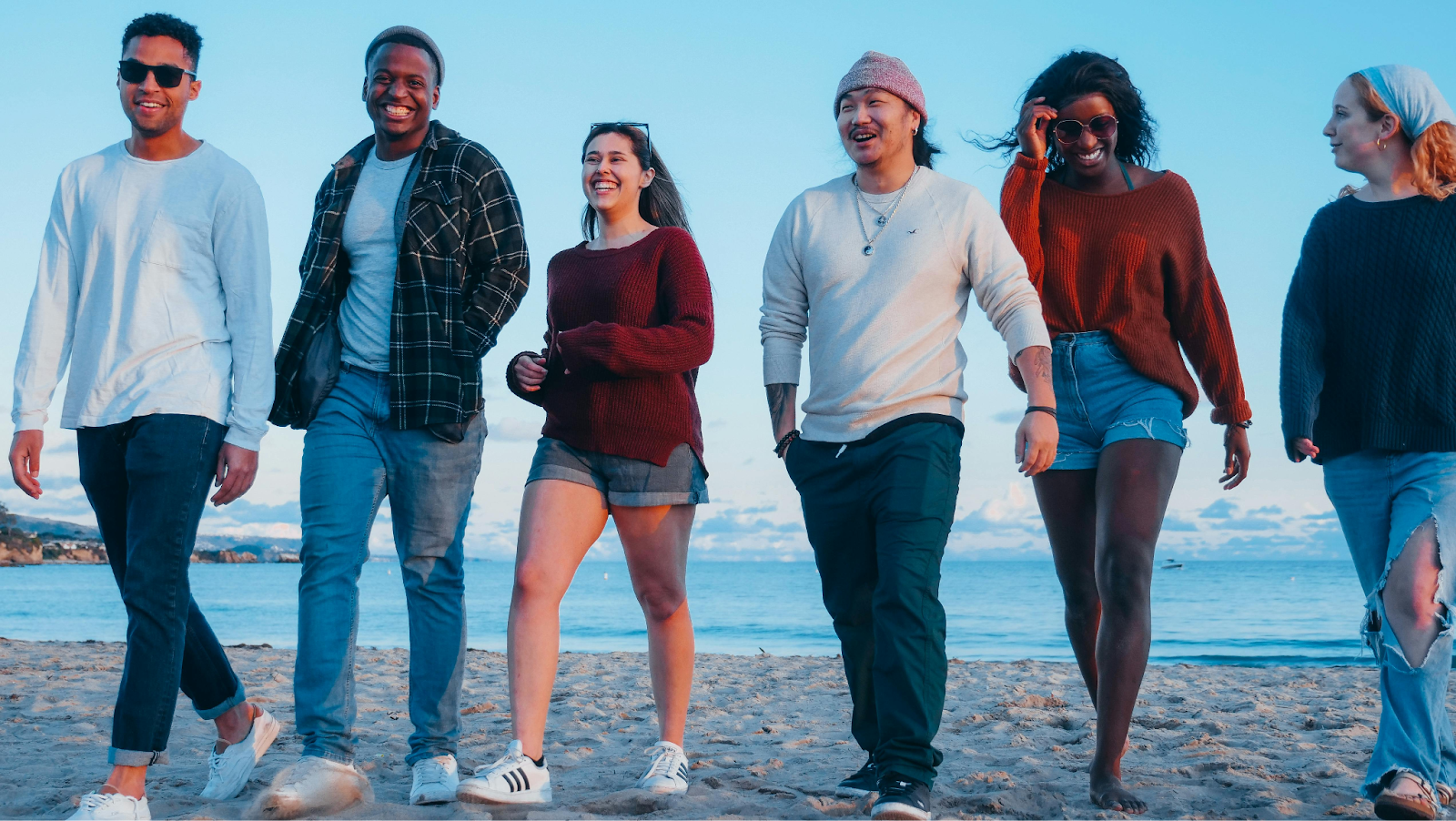 A diverse group of six friends in casual autumn clothing walk and laugh together along a sandy beach. The ocean and a pale blue sky meet at the horizon behind them during the "golden hour."
