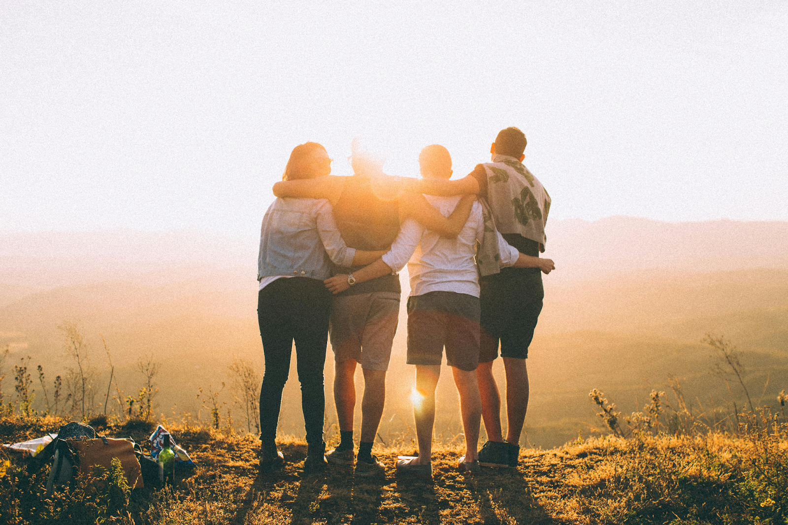 Four friends stand with their arms around each other's shoulders, viewed from behind, as they look out over a vast, hazy valley at sunset. The sun glows brightly between them, creating a warm, golden rim light.