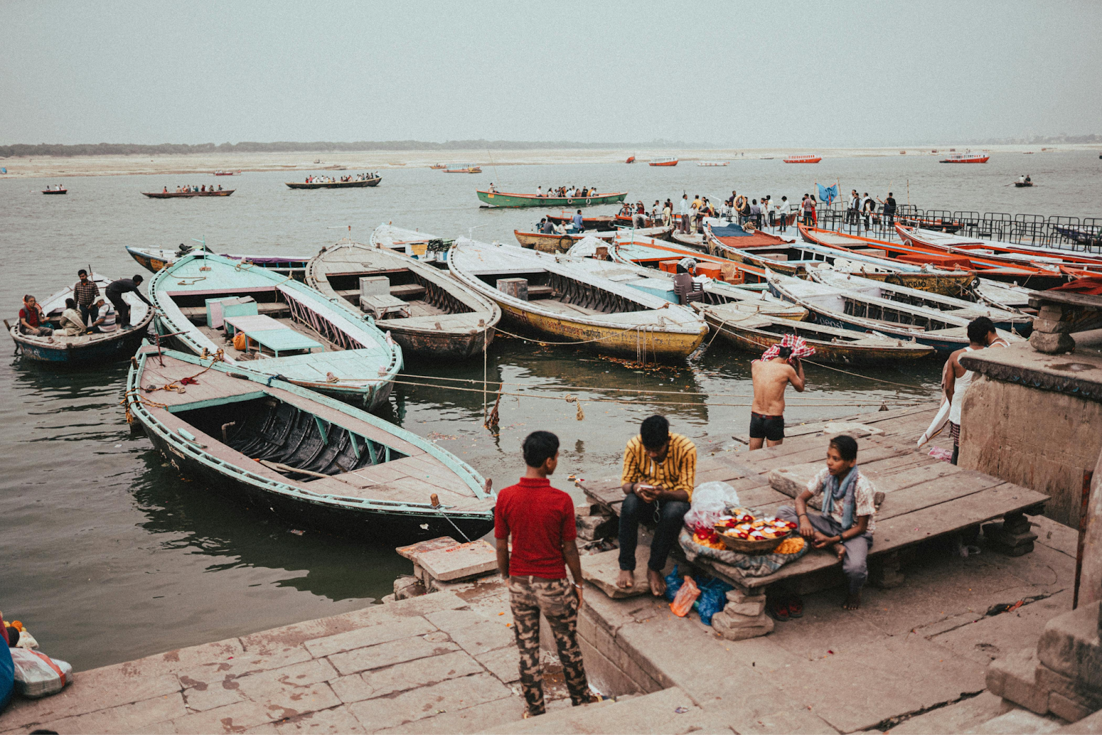 Several colorful wooden boats are docked along a stone ghat by a wide river. On the shore, people are gathered, and a street vendor sits behind a small table of goods.