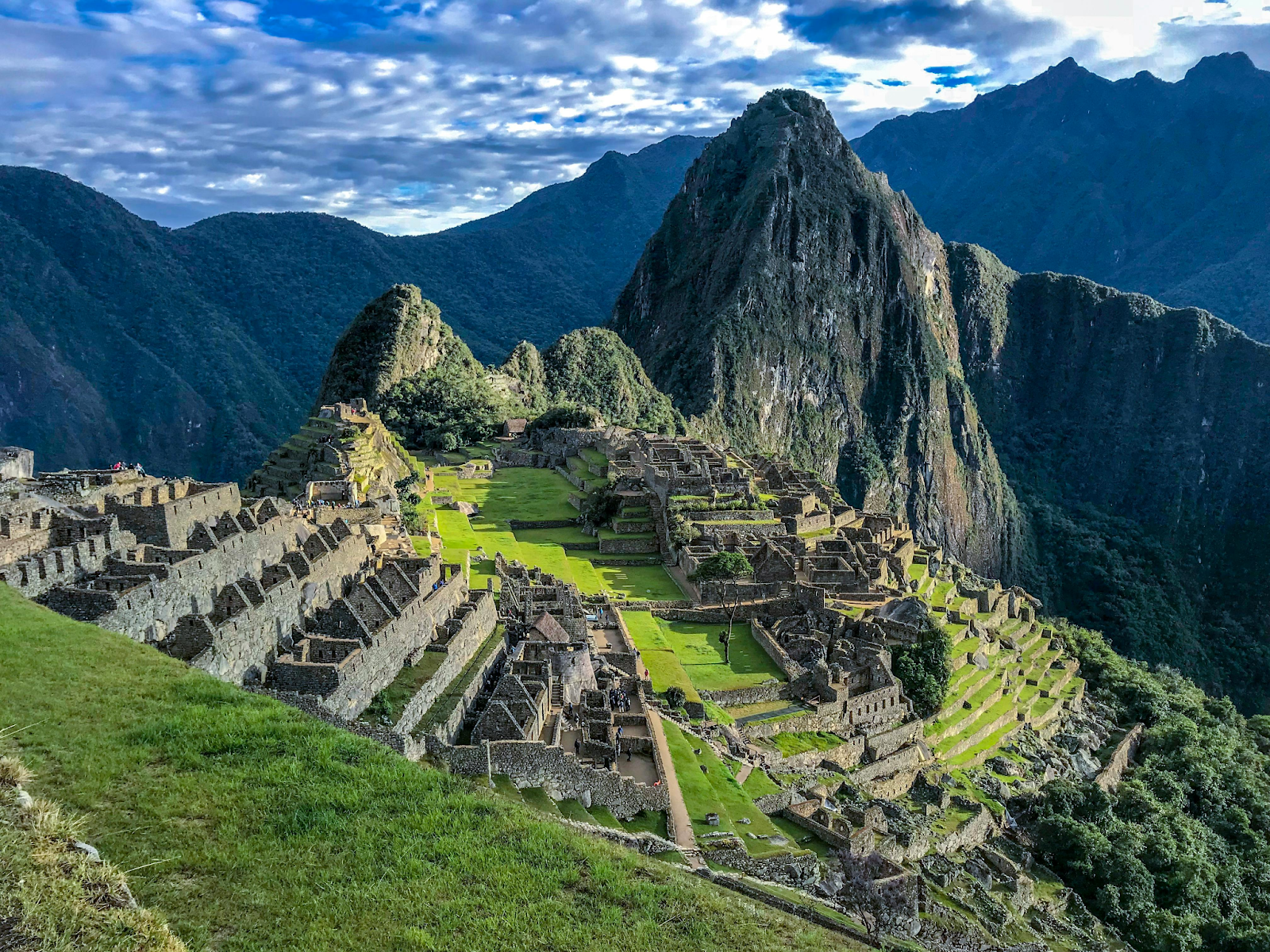 A high-angle view of the ancient stone ruins of Machu Picchu, nestled on a lush green mountain ridge with a steep, pointed peak in the background under a cloudy sky.